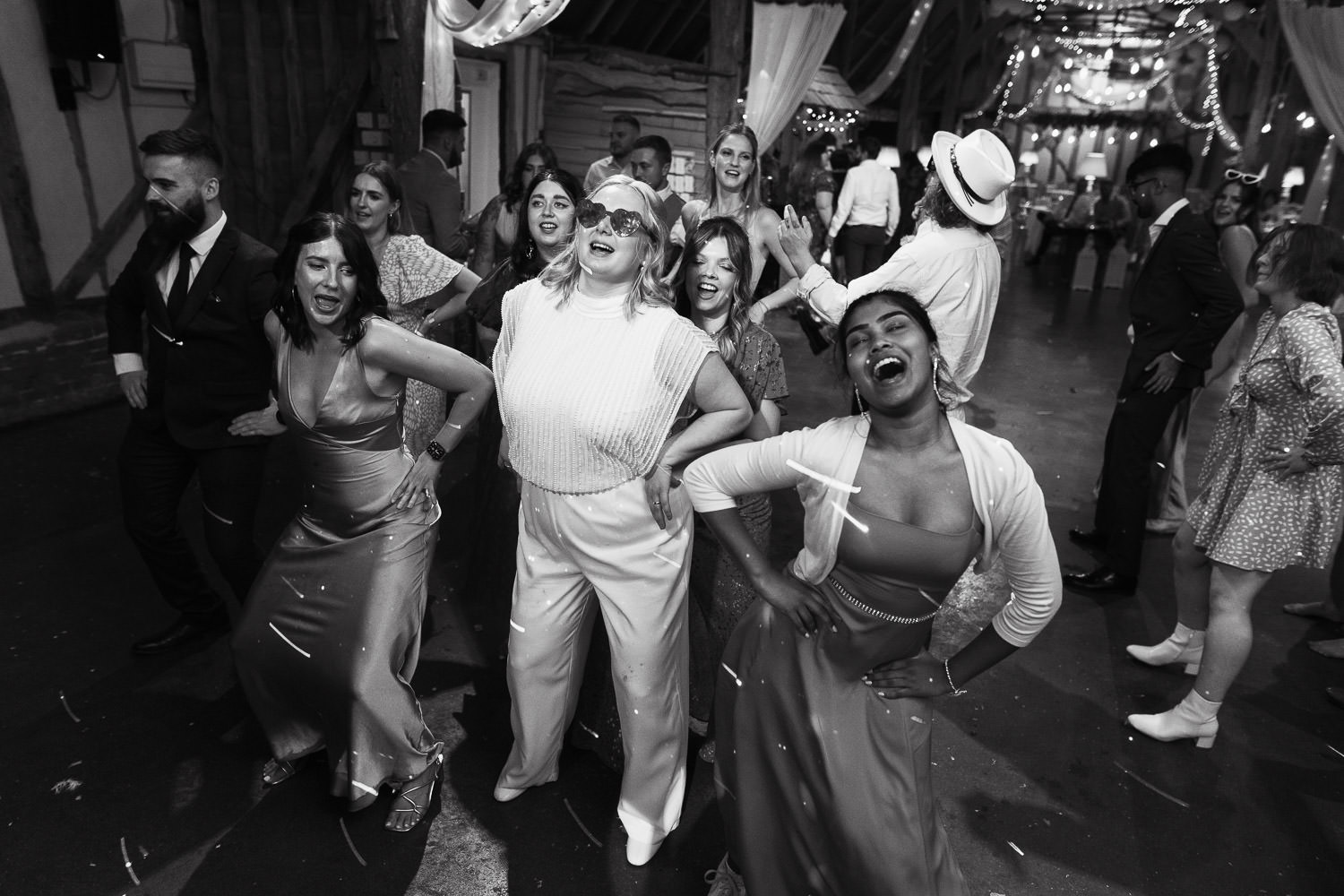 A full dancefloor, people dancing with their hands on their hips. A bride in the middle has pink heart shaped glasses on. Alpheton Hall Barns.