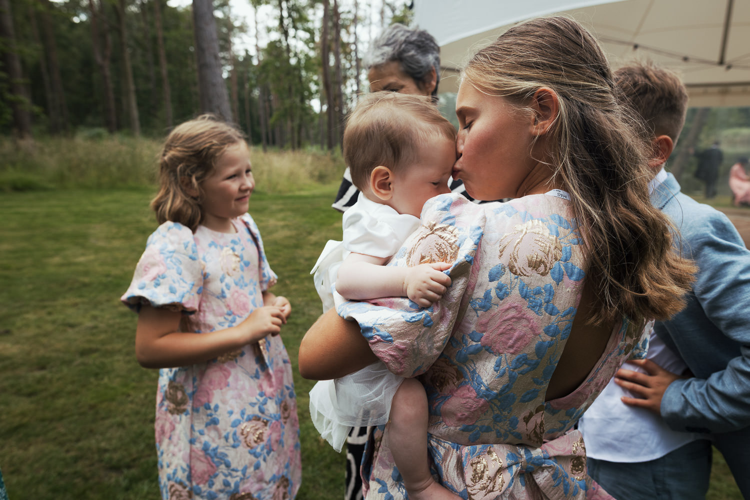 Young girl wearing Marks & Spencer Floral Jacquard Print puff sleeve dress kissing a baby on the forehead. Candid wedding photos of children.