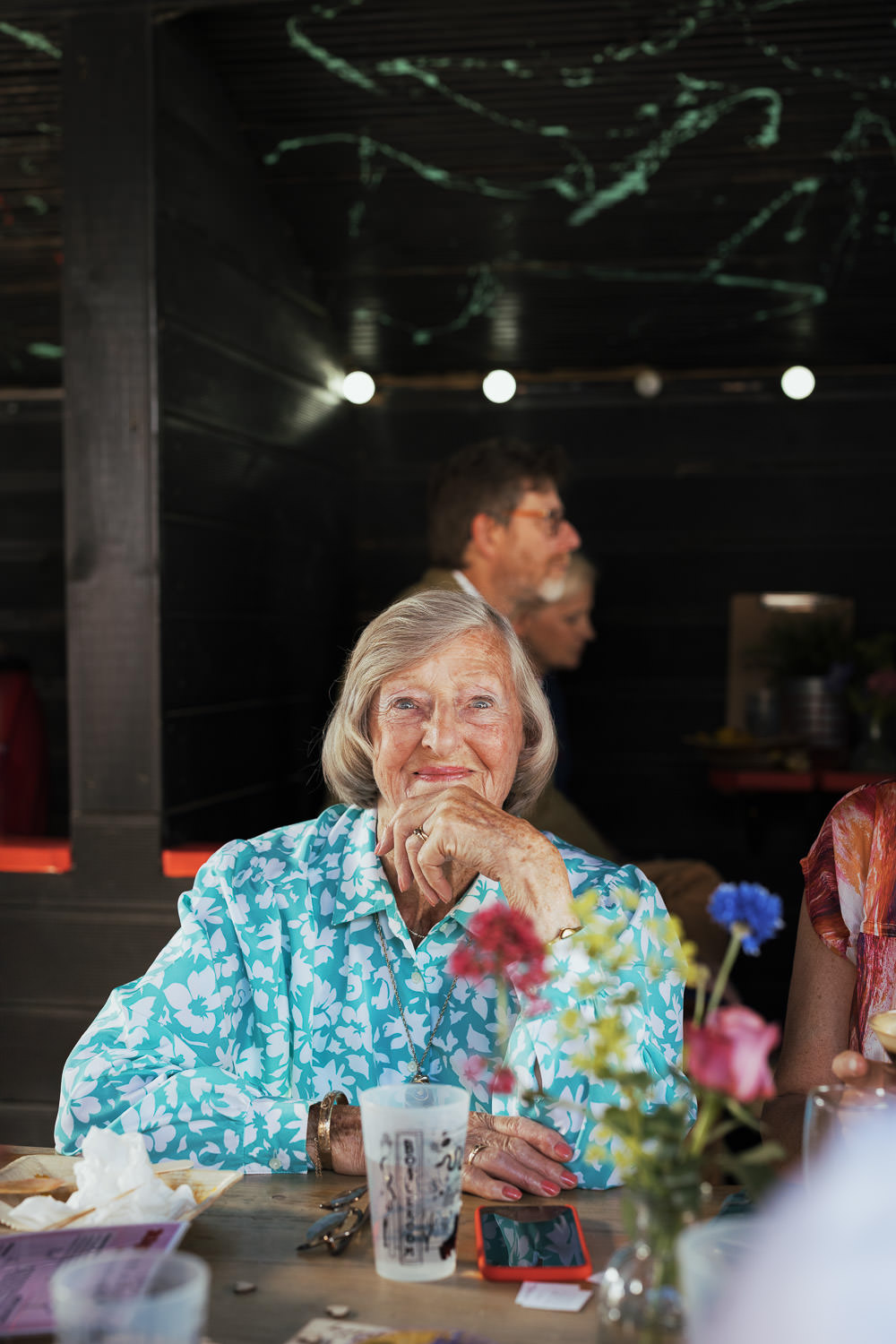 Older woman with fair hair in a bob, wearing a green blue and white floral dress. Sat at a table with wedding guests, smiling at the camera. A natural wedding guest portrait.