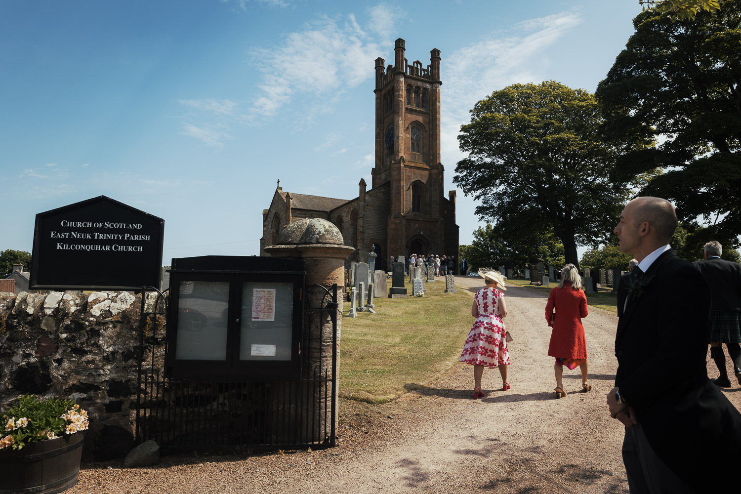 Usher standing my gate of the church. The sign says Church of Scotland East Neuk Trinity Parish Kilconquhar Church. Guests walk up the path.