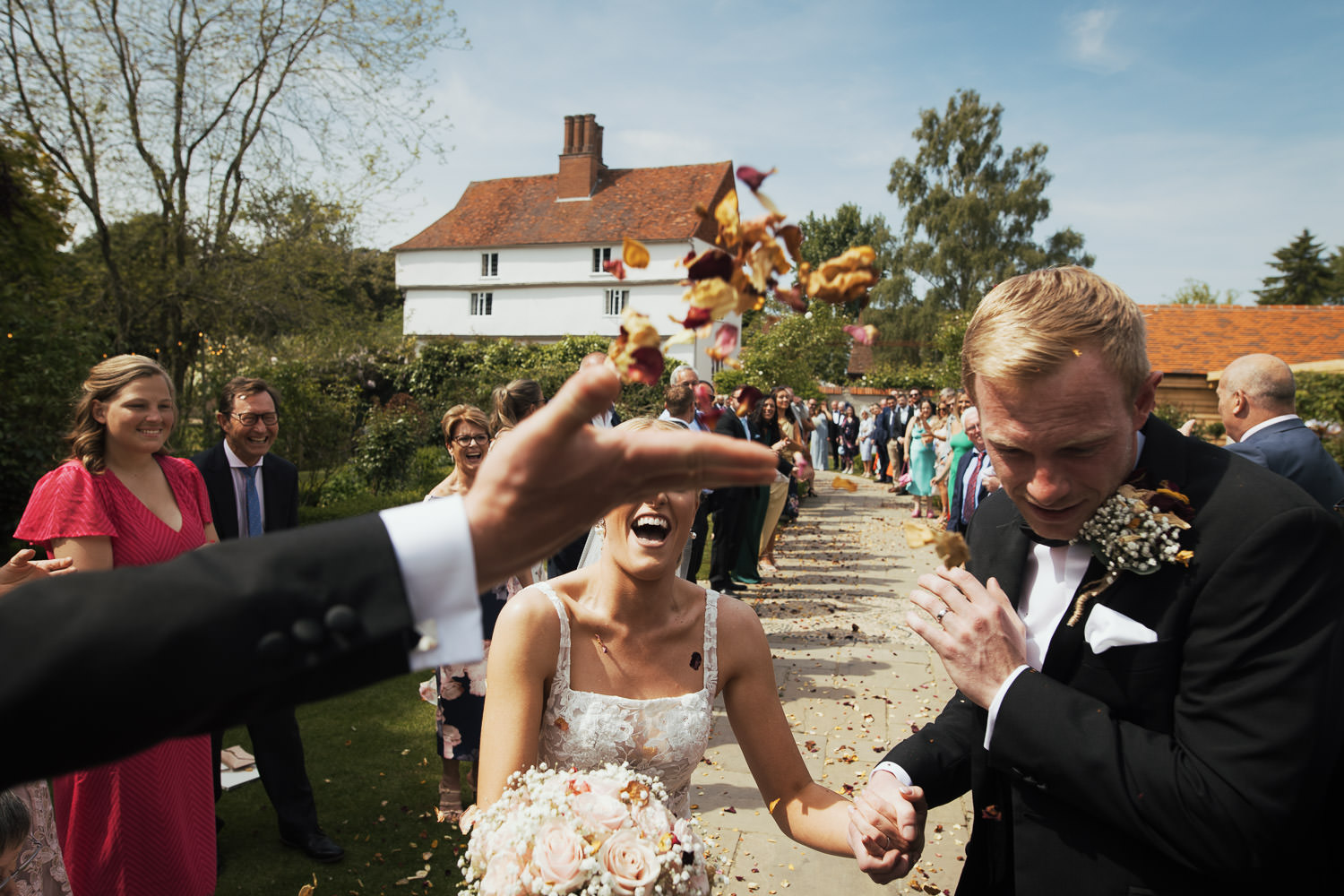 Newly married couple walking through confetti tunnel. Hand throwing confetti. Black tie suit from The Groom’s Room in Colchester. Dress called Jack by Made With Love from Halo + Wren bridal. At Houchins venue, farmhouse and blue skies behind them.