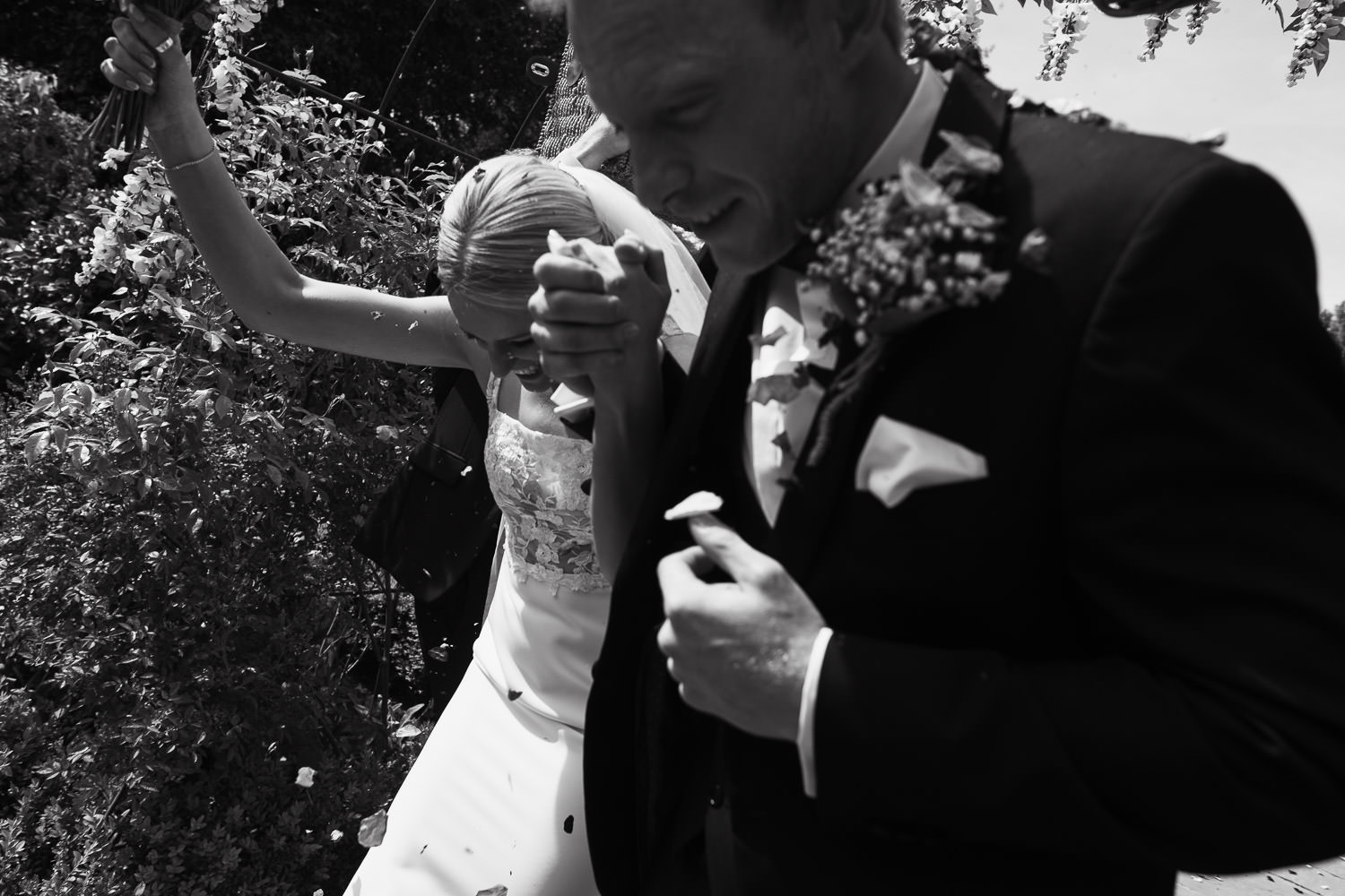 Essex wedding photographer at Houchins venue near Colchester.  Bride and groom passing by the camera after a confetti tunnel. Looking very happy. The groom is wearing black tie tuxedo from The Groom's Room in Colchester. The bride is wearing a dress called Jack by Made With Love Bridal from the Halo + Wren bridal shop.