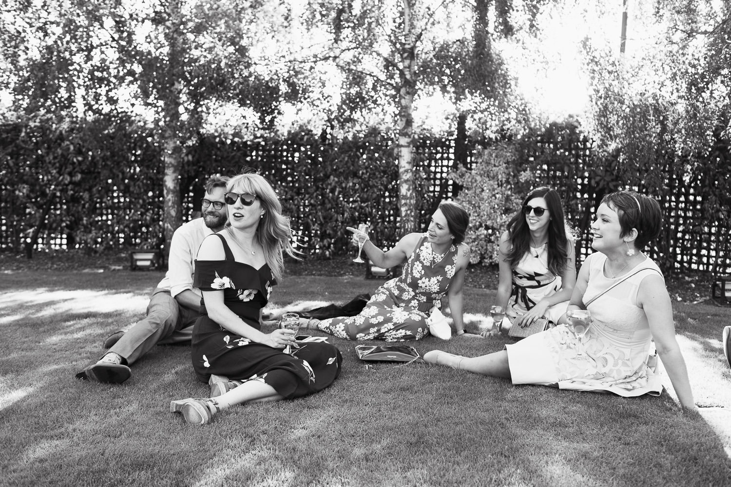 Five wedding guests sat on the grass on a warm day at Crondon Park venue near Chelmsford. Woman with long hair is wearing Warehouse, BUTTERCUP COLD SHOULDER DRESS. Woman in the middle is pointing, wearing a floral dress. Person on right with shorter hair has a pale dress.