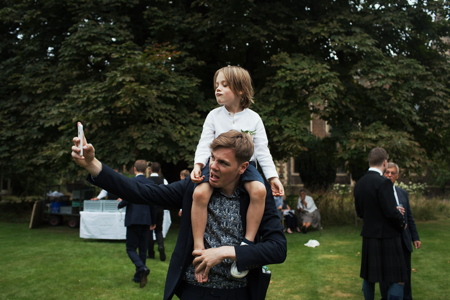 Boy on the shoulders of a young man, pulling faces taking a selfie, in the garden of St Johns College Cambridge wedding.