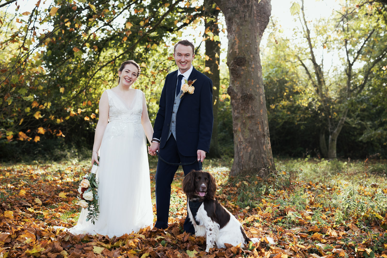 Couple portrait in the trees at Essex venue Houchins in the autumn. The bride is wearing Jenny Packham and the groom in a dark blue suit. They are with their dog, a springer spaniel.