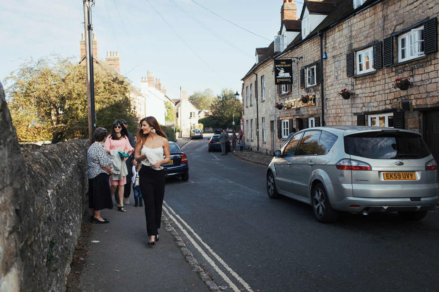 Wedding guests arriving at St Andrew's Church in Headington. The White Hart pub in the right. A woman is wearing white strappy top and black trousers as she walks up the path.