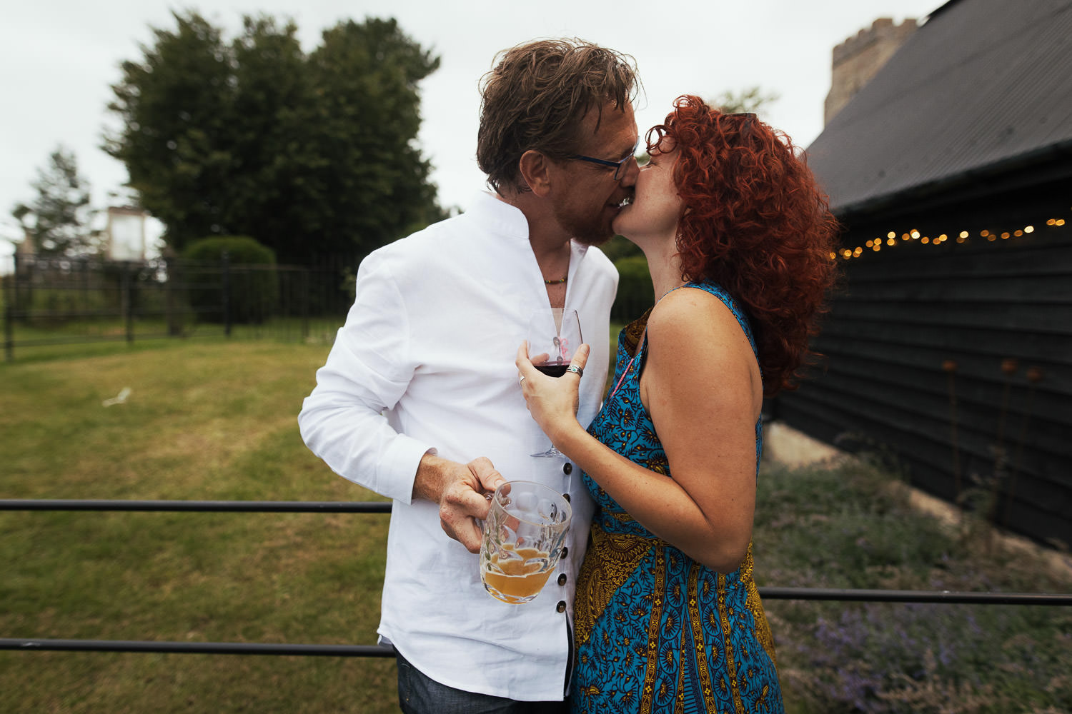 A couple of wedding guests at a wedding outside the Alpheton Hall Barn. They are kissing. She's holding glass of red wine, he's holding a Dimpled Pint Glass of beer.