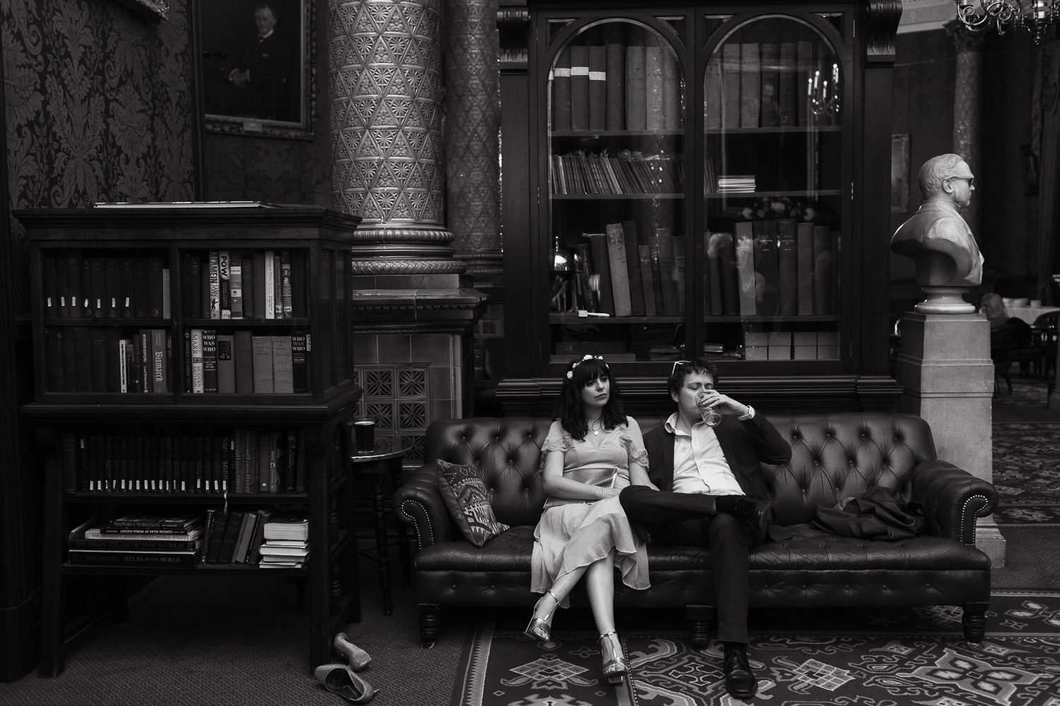 Couple sat on a sofa at a wedding reception in the smoking room of the National Liberal Club. The bust of Joseph Firth Bottomley Firth on the right. A bookcase on the left.