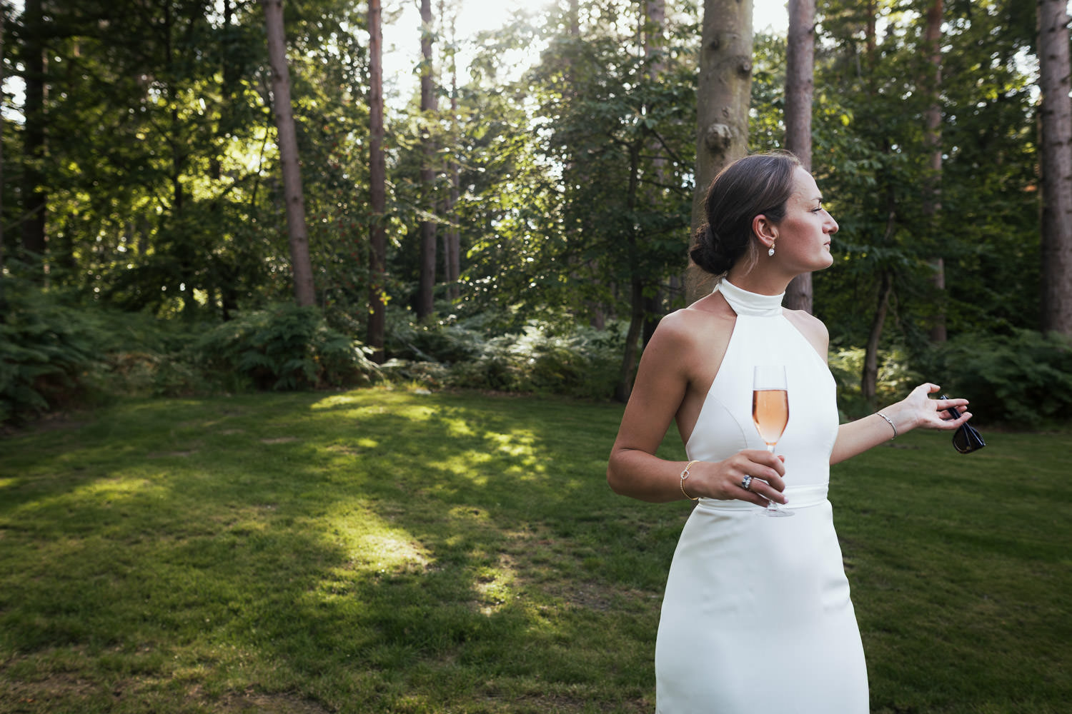 Candid bridal portrait in the trees with dappled sun. Bride wearing a halter neck dress by Katrine Mogensen Bridal Couture and holding sunglasses.