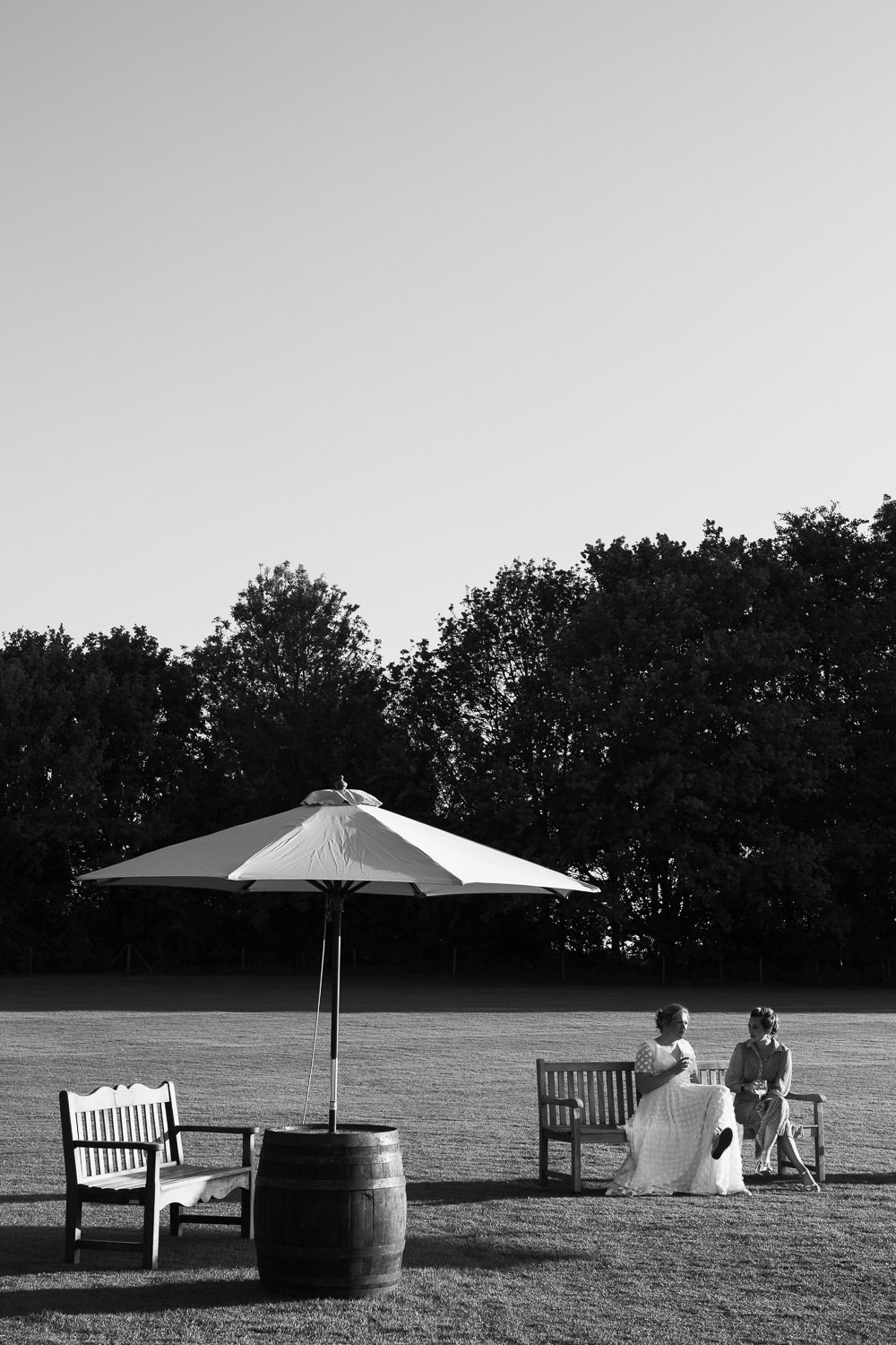 Bride and bridesmaid sat on a bench in the sunshine, drinking wine. Lindsell.