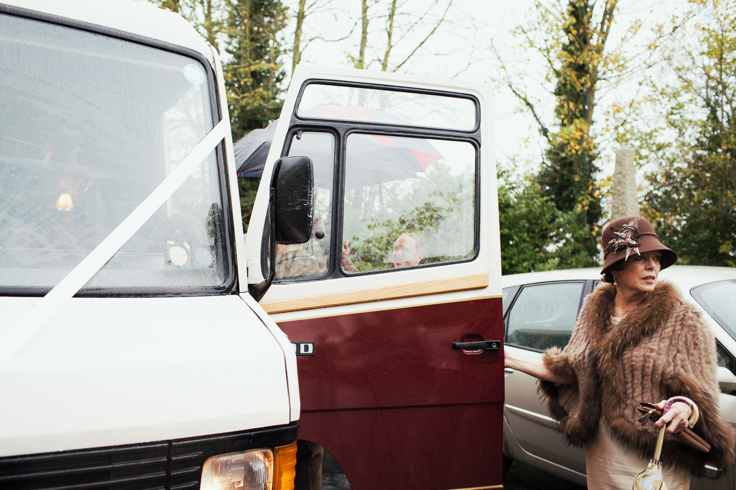 Mother of the groom in a brown furry shawl and hat is outside in the rain. About to get aboard a coach.