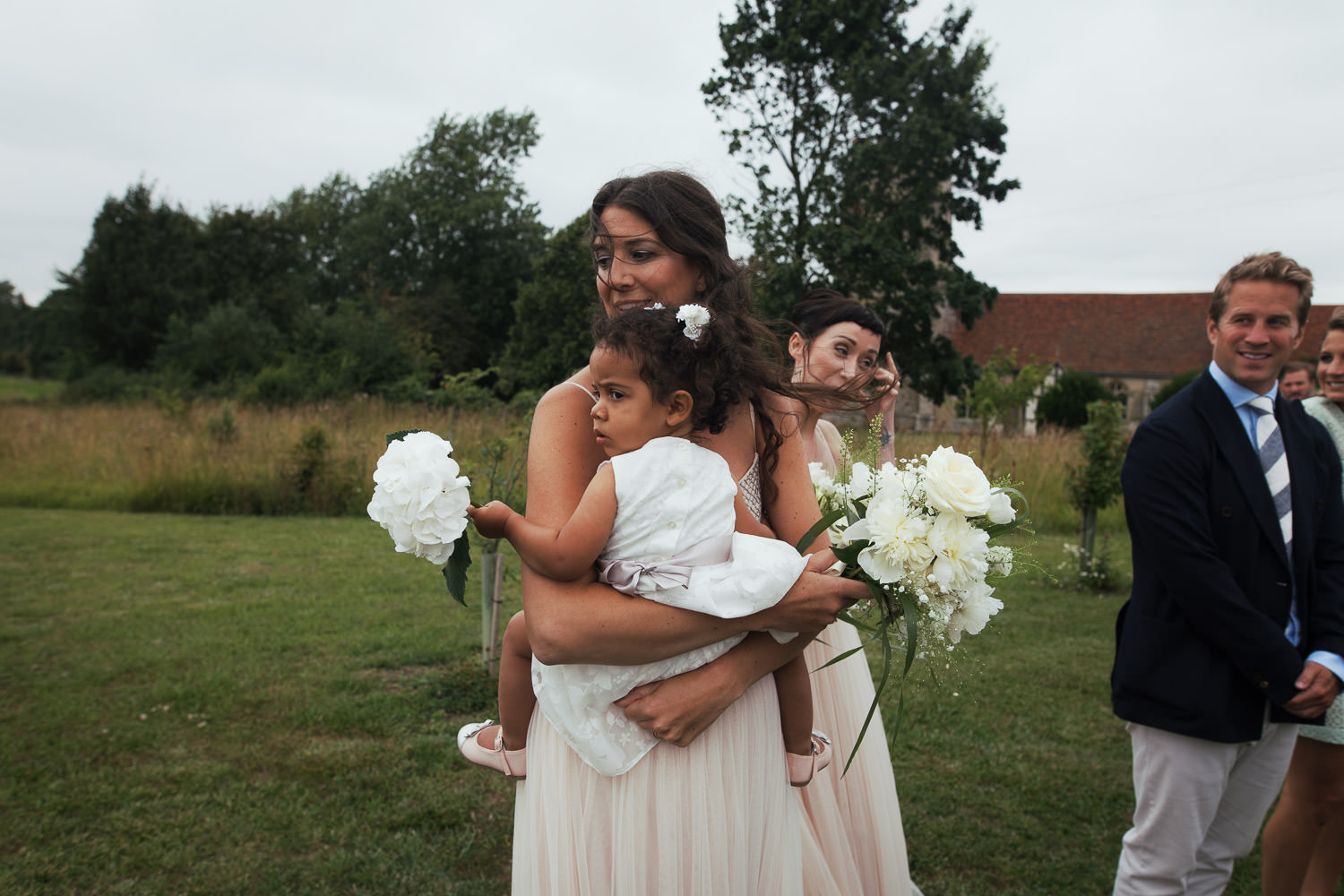 A bridesmaid in pink holds her young daughter the flower girl. Natural wedding photography at Alpheton Hall Barns in Suffolk.