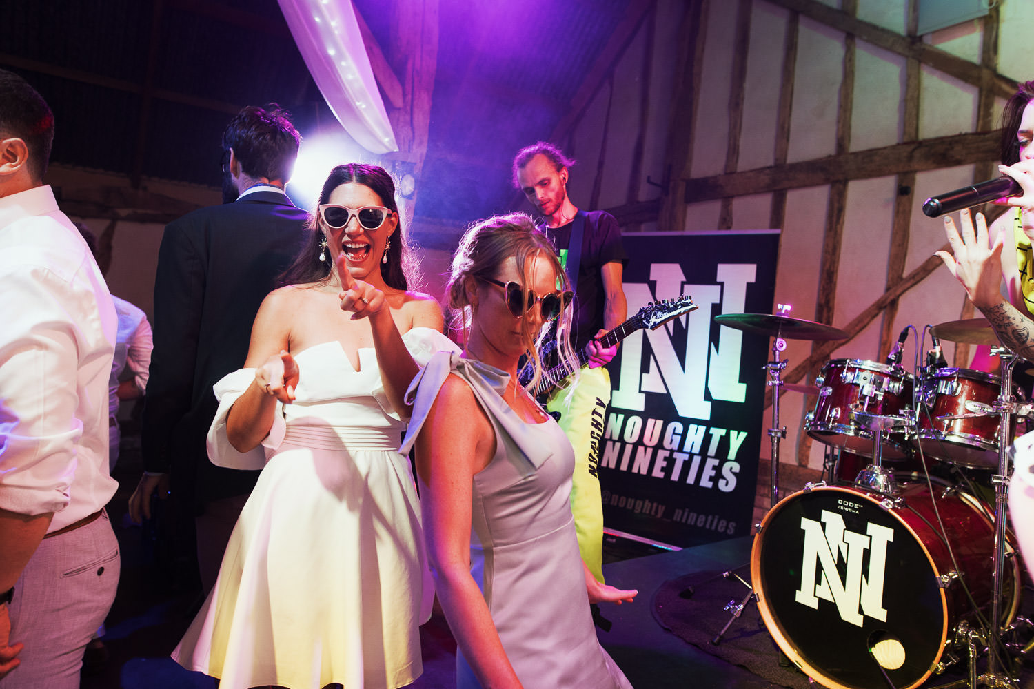 A bride at her wedding reception wearing ASOS Trendyol bandeau sleeve mini dress in white and sunglasses. With her bridesmaid. They are dancing to the band Nought Nineties at Alpheton Hall Barns. Taken by Essex wedding photographer.