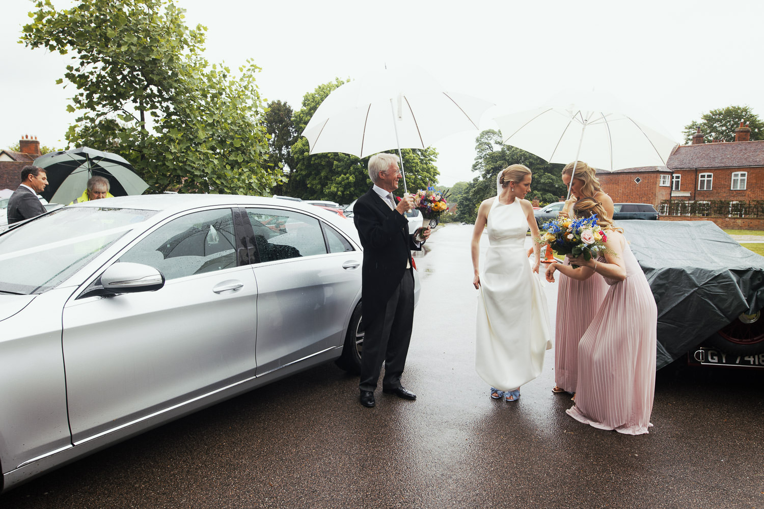 A bride wearing Jesus Peiro 260 dress from Miss Bush is outside the church in Danbury Essex. It is raining and her dad and bridesmaid hold an umbrella. Documentary style Essex wedding photographer.