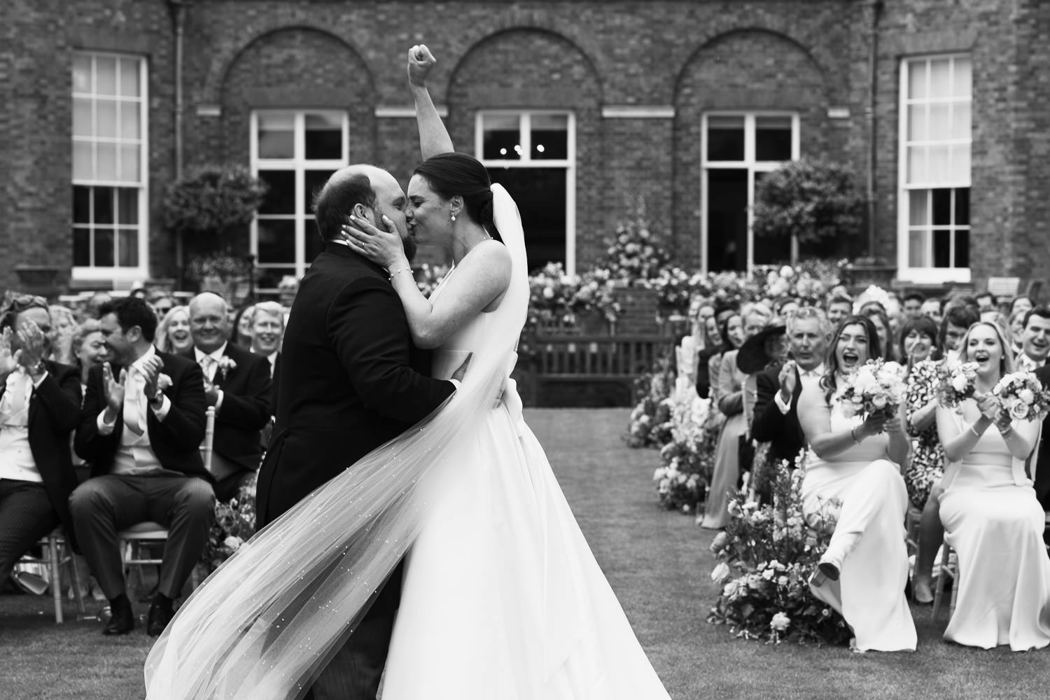 Bride punches the air with happiness and kisses her husband during wedding ceremony at the Jockey Club Rooms in Newmarket. They are outside on the lawn and guests are clapping. 101 High St, Newmarket CB8 8JL