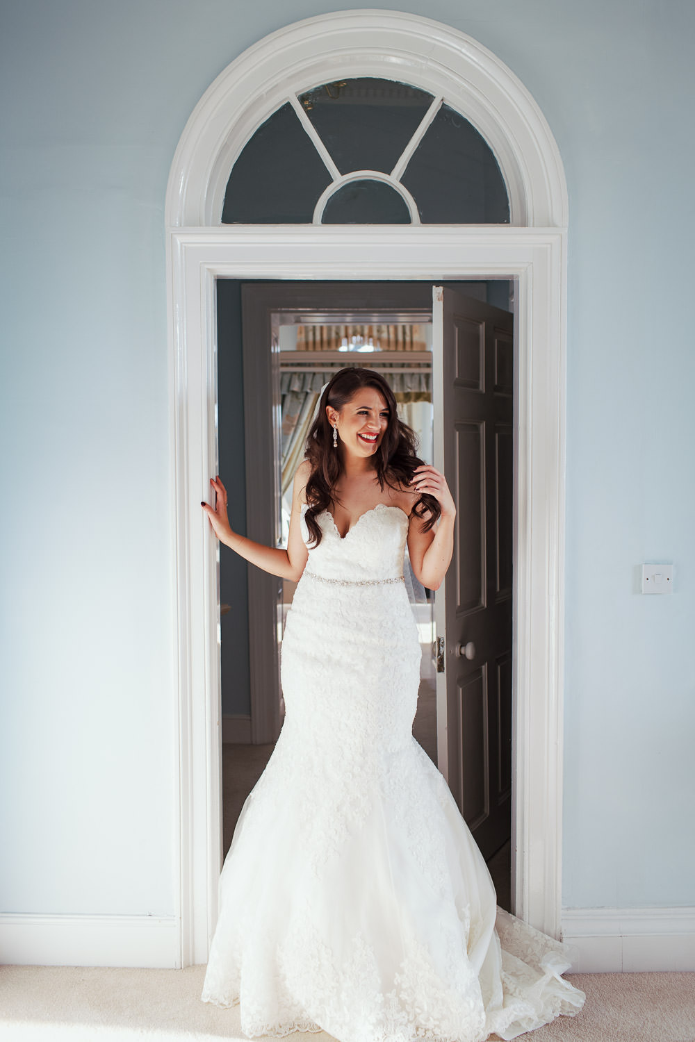 Documentary wedding photo: Bride with long dark hair and red lipstick, smiling in the doorway of the Queen's Apartment at Gosfield Hall venue, Essex. She wears a Mori Lee lace mermaid wedding dress (style 2713).