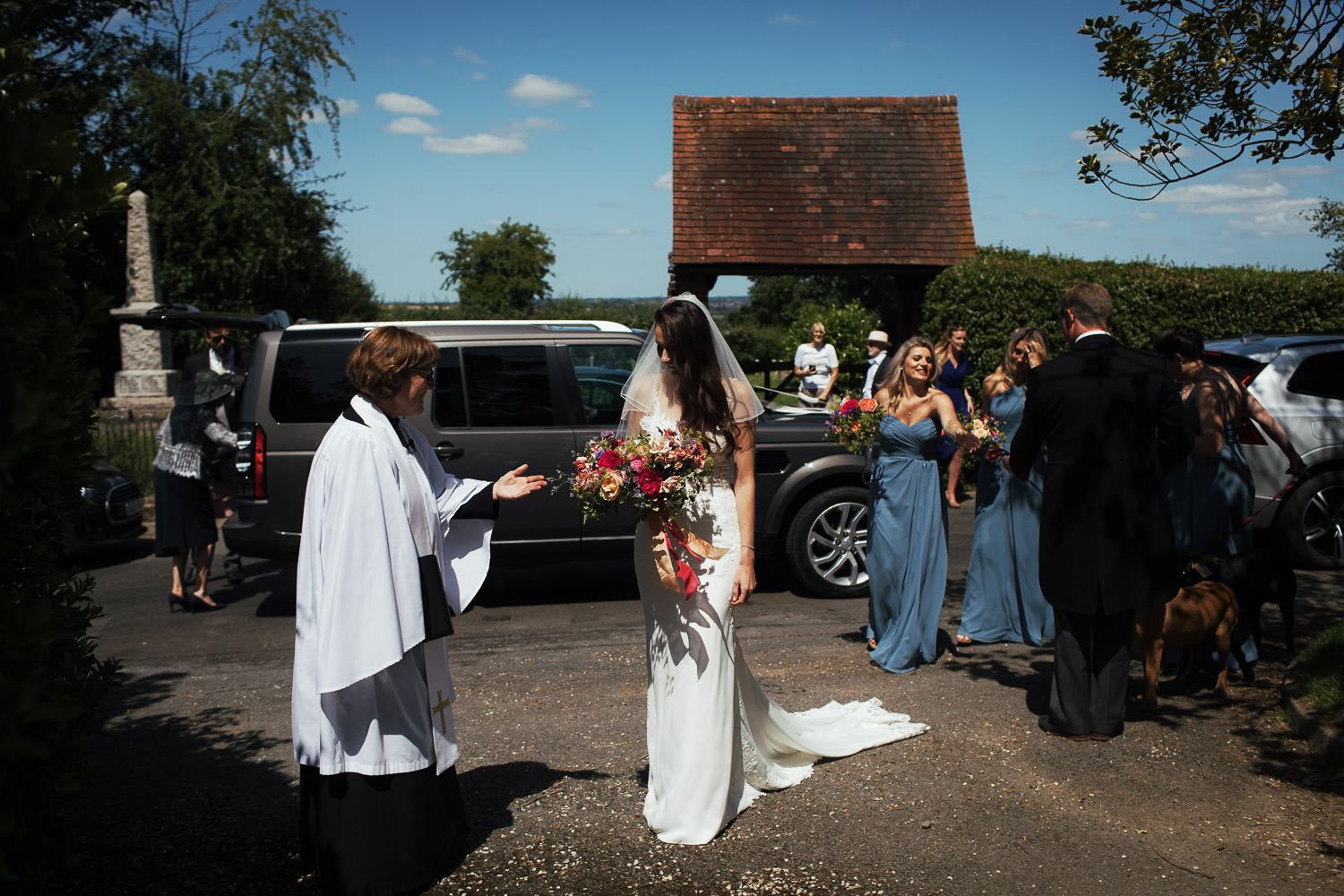 Purleigh Church Wedding Photography: Reverend Julie Willmot welcomes a bride in a Stella York 6648 gown from The Wedding Shop Colchester. Bridesmaids in blue dresses stand behind her against a blue sky. Captured by Tracy, an Essex documentary wedding photographer.