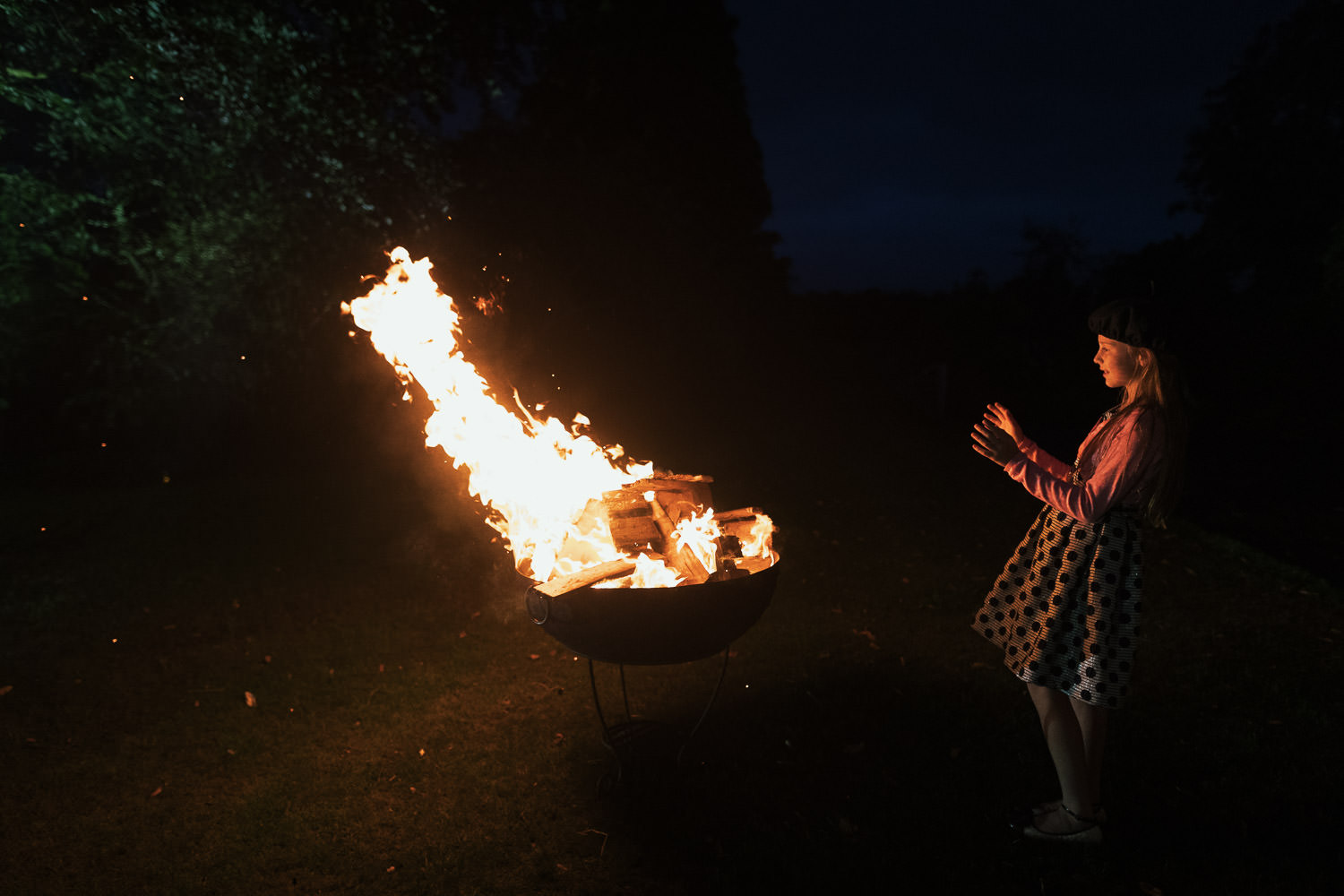 A girl in a spotty dress and pink cardigan warms hands by a burning fire pit.