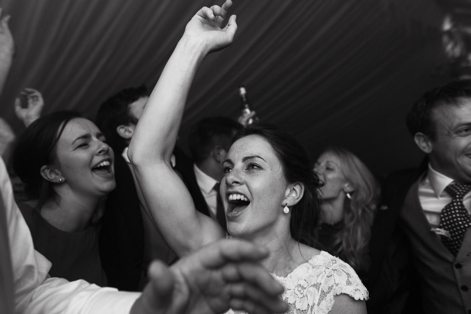 A crowded wedding dance floor captures the bride singing and dancing with hand in the air, surrounded by guests enjoying live music from Monstaball. Bride wears Essense of Australia style D2009 patterned lace dress from Shropshire Country Brides.