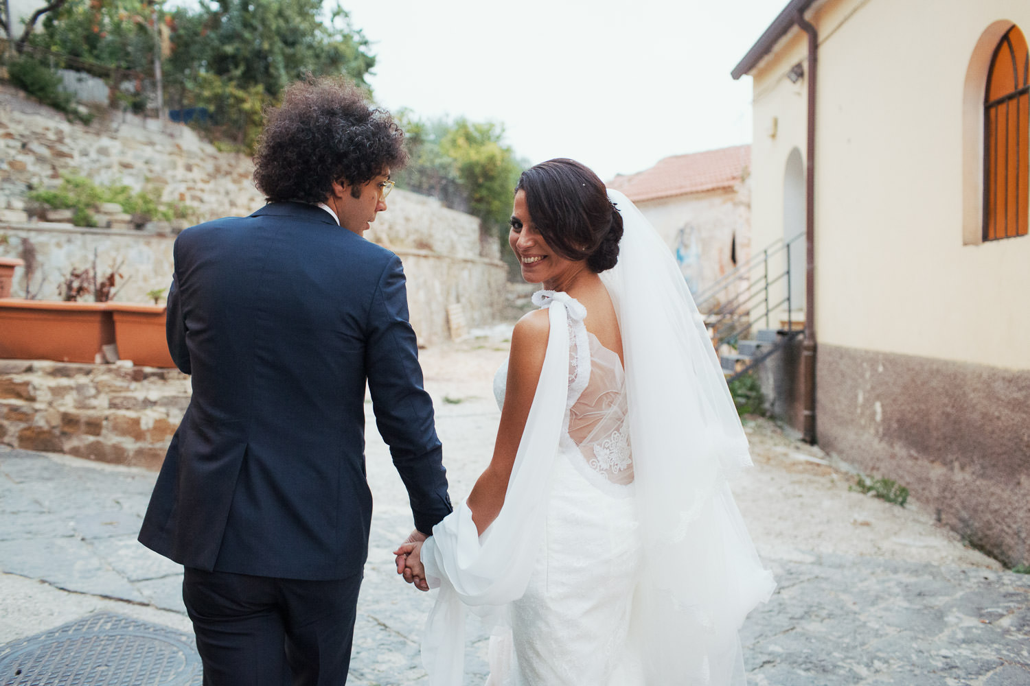 Bride and groom walking along holding hands. Bride looks over her shoulder at the camera.