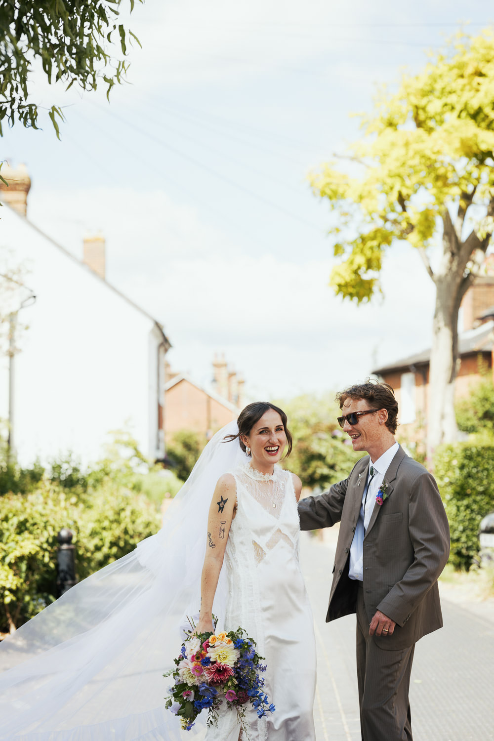 Bride in AllSaints Lace Mila Midi Dress with high neckline, ruffle detail, and lace hem, laughing with groom in beige cowboy-core suit on Stoke Fields, Guildford. Flowers by Holly Tree.