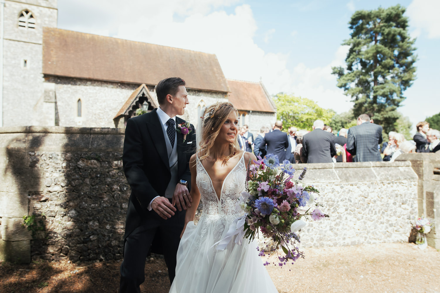 Newly married couple standing outside St Peter and St Paul's church in Shiplake. Wedding guests behind the wall. Documentary style wedding photography.