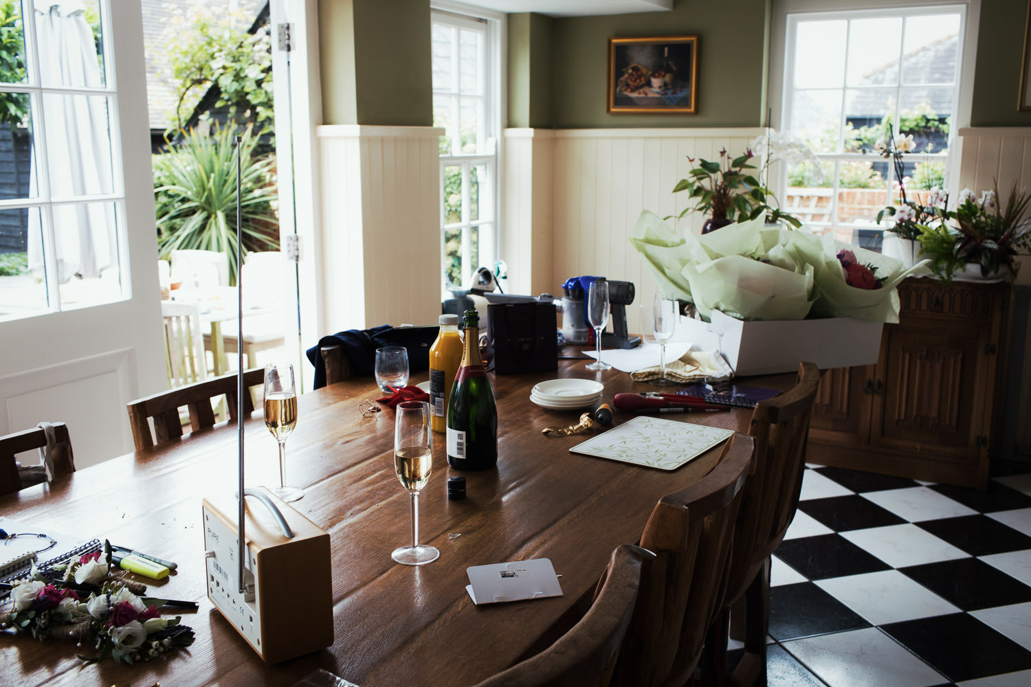 At home before a wedding. There are half drunk glasses of Bollinger, a radio, and flowers on a dining room table. A wedding in Great Totham. Natural wedding photography.