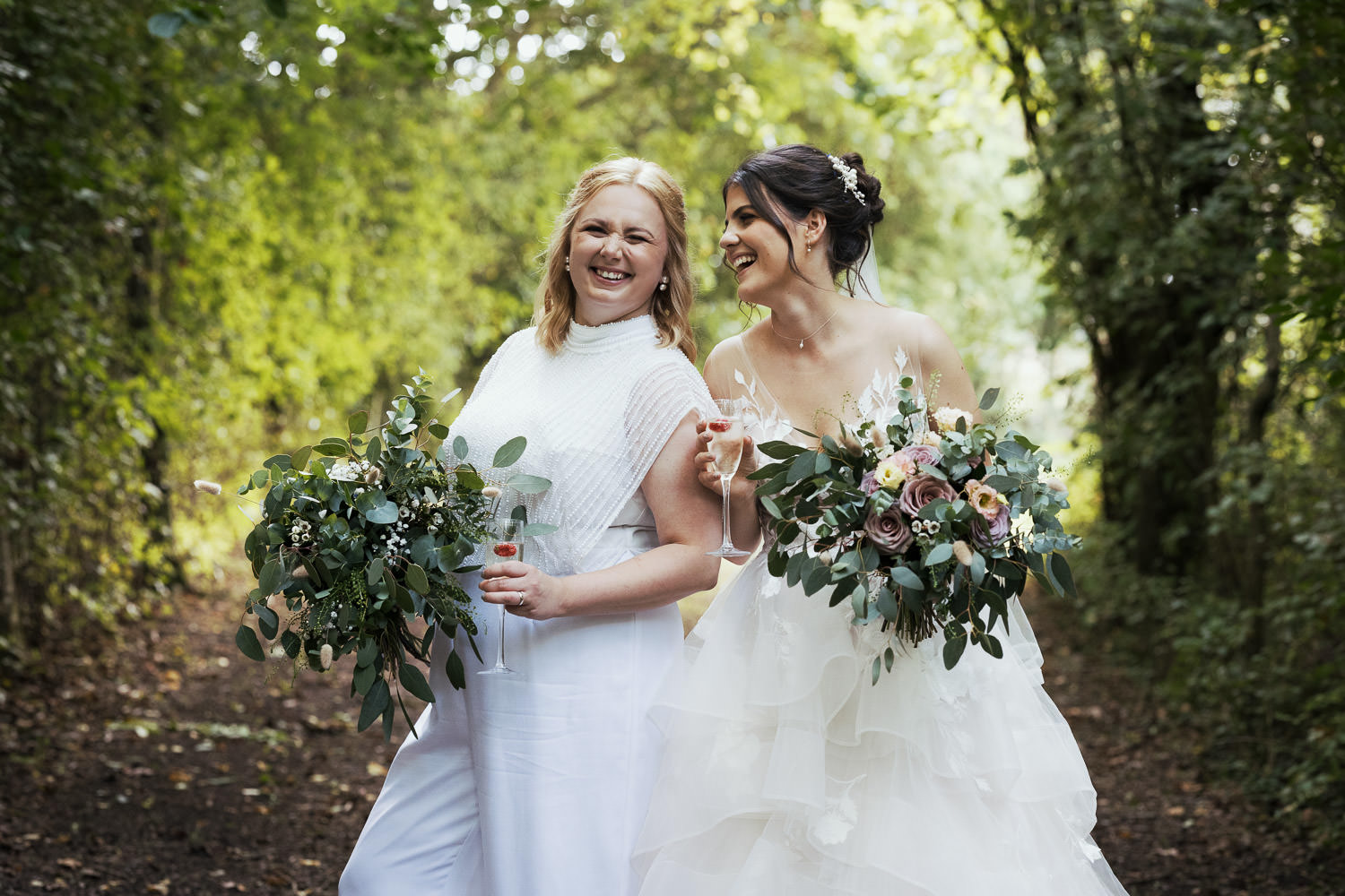 Two brides in the lane at Alpheton on their wedding day. Holding flowers from Wild + Mae. The bride with brown hair is wearing a dress. The Wtoo by Watters Montgomery 12716. The bride with blonde hair is wearing the Joanna Hope Beaded Bridal Jumpsuit.