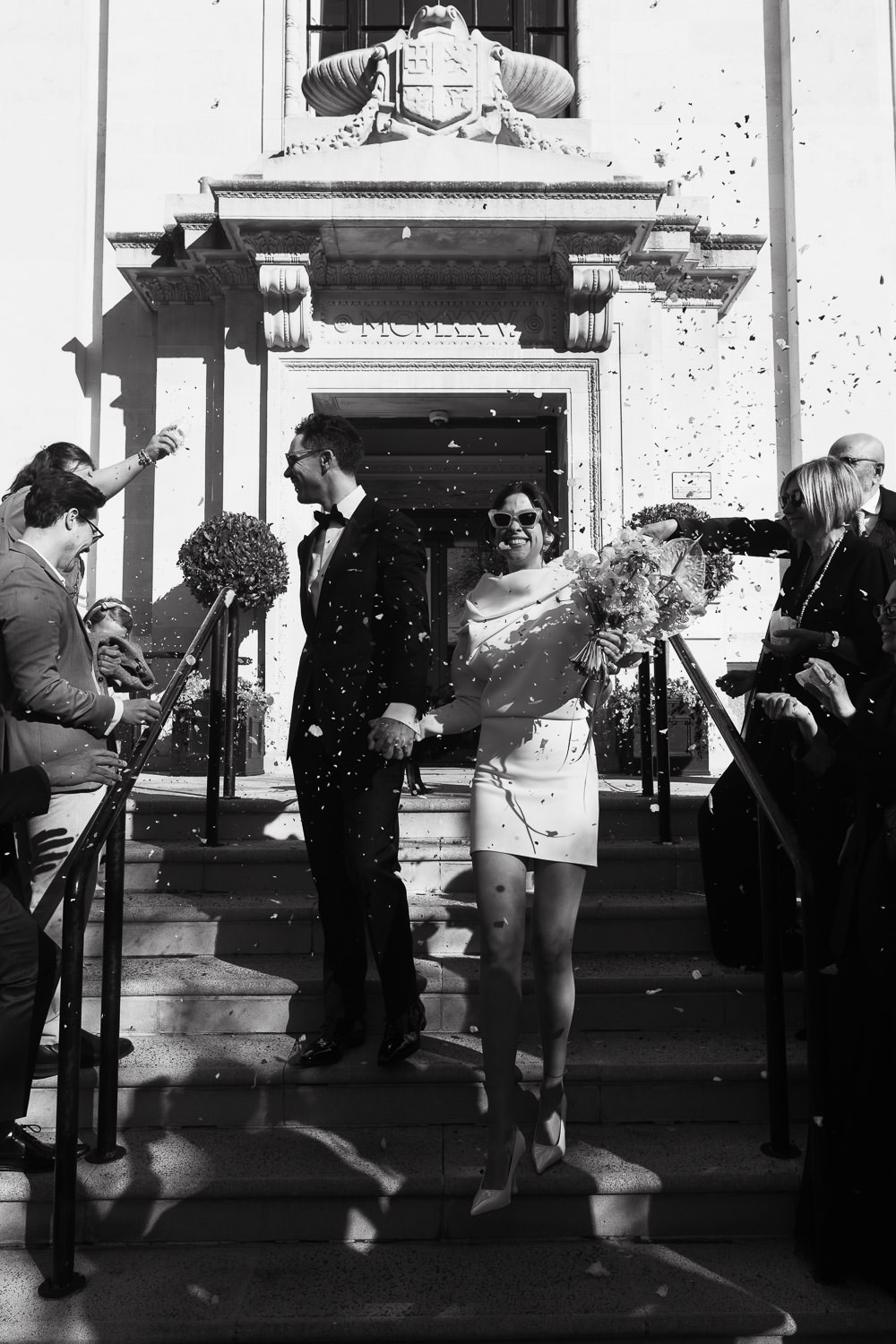 Couple walking down the steps of the Islington Town Hall in the sunshine. Family and friends throw confetti at them.