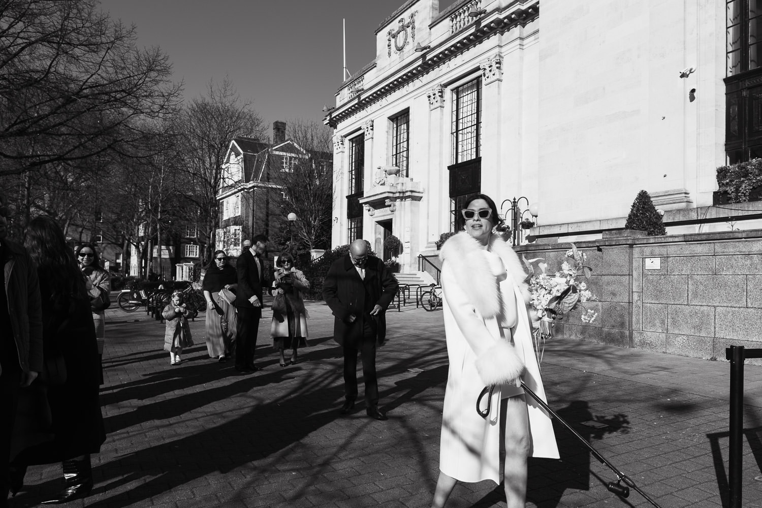 Islington Town Hall Wedding: Bride in a short dress, coat and sunglasses walking away from the town hall holding the dog lead.