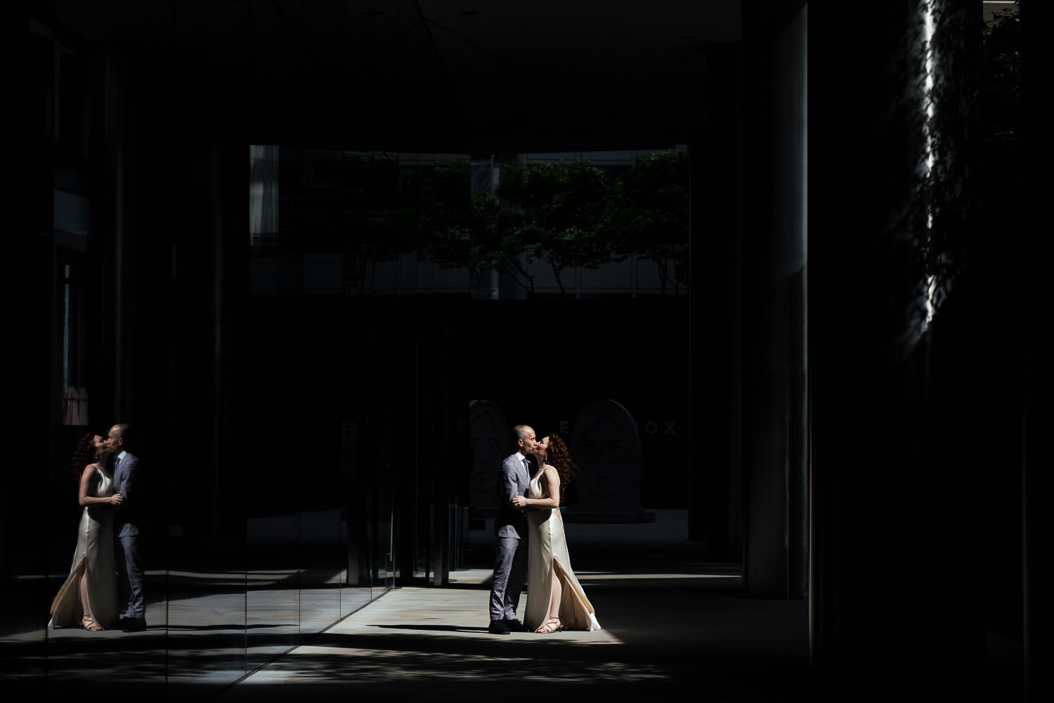 Newly married couple share a kiss on Clerks Place in Bishopsgate after their St Ethelburga's wedding. The bride wears a silk satin bias-cut wedding dress with spaghetti straps. Shadows from the stone pillars fall around them, and their reflection is visible in the glass facade of 100 Bishopsgate.