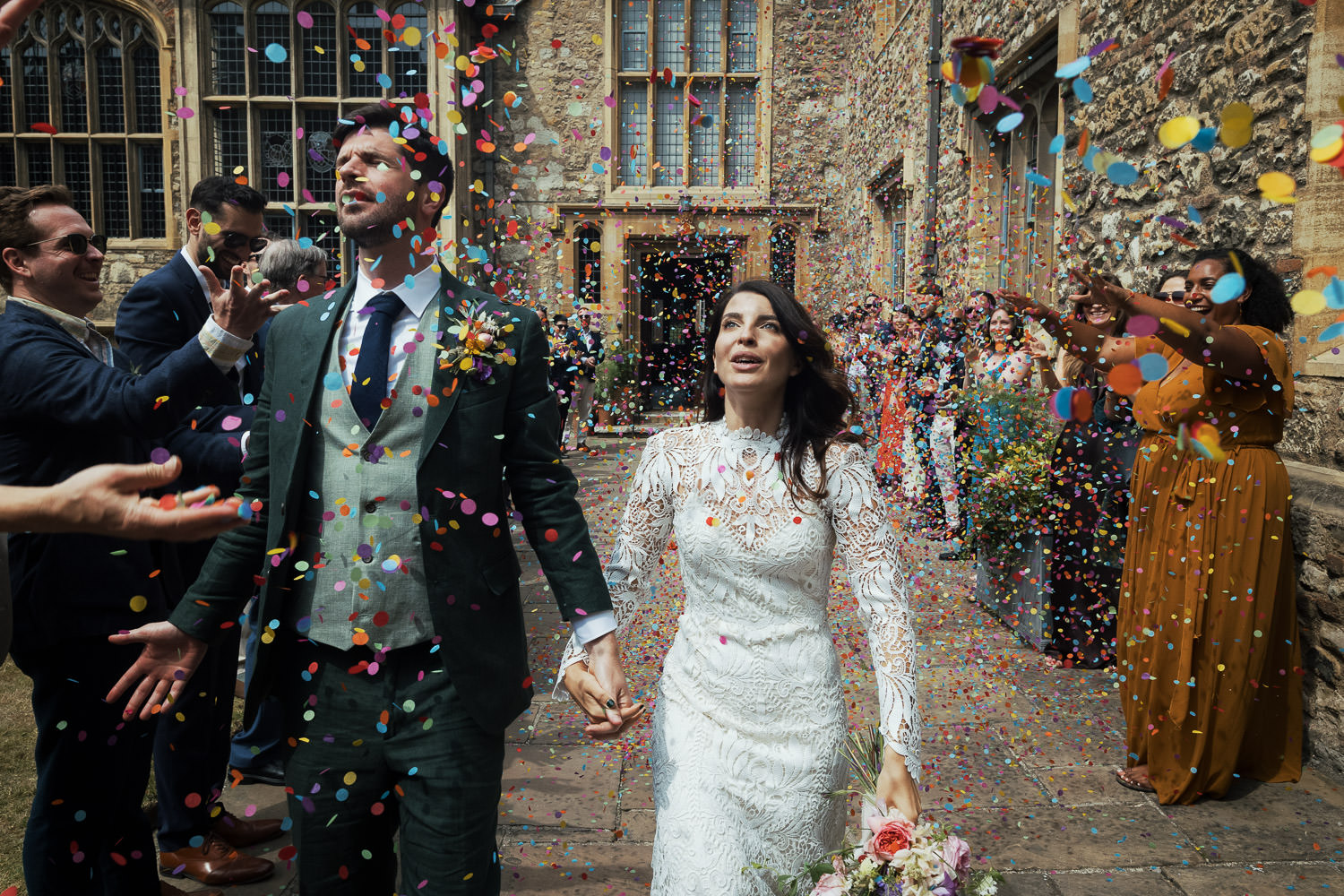 The Charterhouse monastery wedding venue, Clerkenwell. Bride and groom walk through their wedding guests who throw colourful confetti. Bride is wearing lace dress called Tori by Catherine Deane.