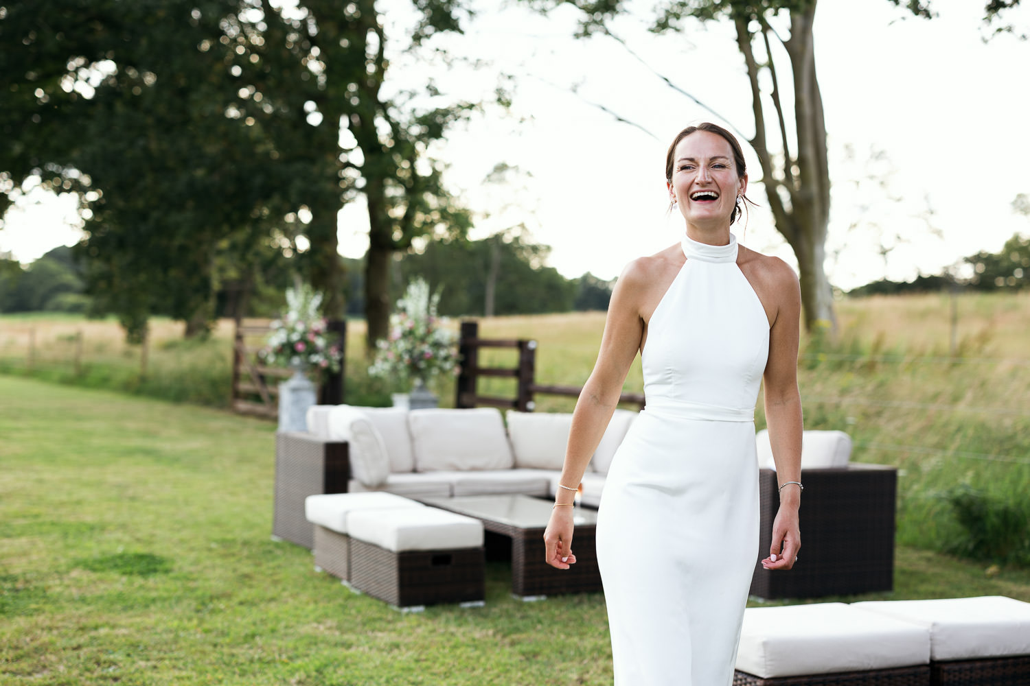 Documentary wedding photographer capturing a natural, candid portrait of a joyful bride in a white halterneck dress by Katrine Mogensen, unposed in a Norfolk meadow.