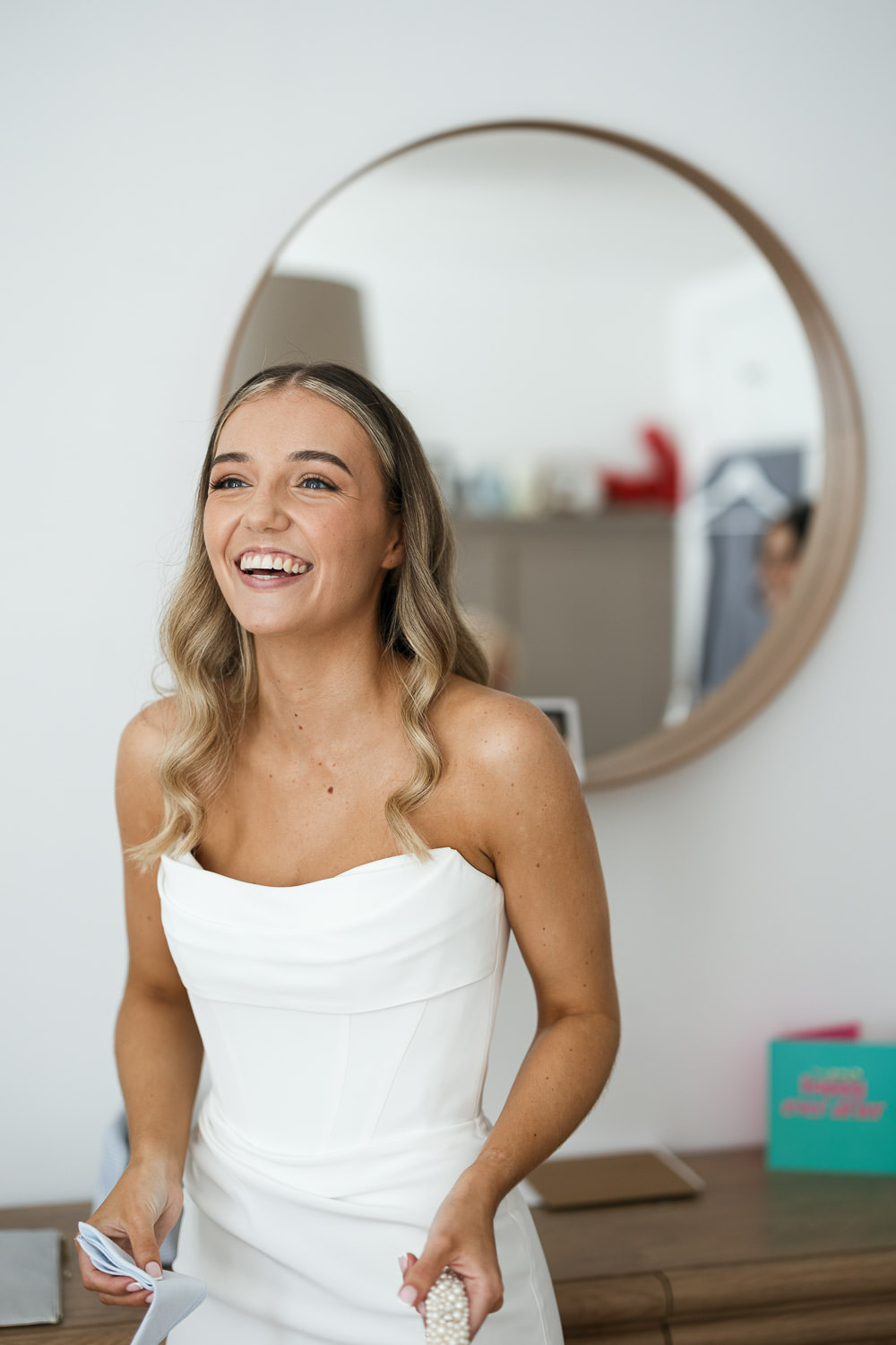 Bride getting ready for wedding in Chelmsford wearing Duke by Chosen By KYHA white wedding dress, long wavy blonde hair, round mirror backdrop.