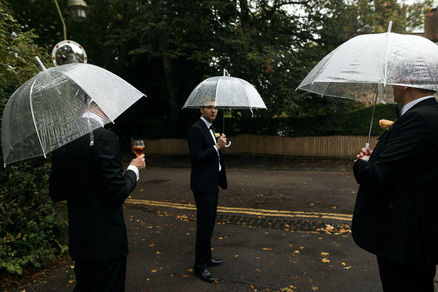 Three groomsmen in black tie, holding clear dome shaped umbrellas in the rain. One man is holding an aperol spritz. They are waiting by the road, Bardwell Road in Oxford.